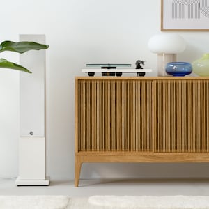 Puede incluir: Un altavoz est&eacute;reo blanco se encuentra junto a un armario de madera con un tocadiscos en la parte superior. El armario tiene un frente de listones y se apoya en cuatro patas. Una l&aacute;mpara blanca con una pantalla blanca y un jarr&oacute;n de vidrio azul est&aacute;n encima del armario. La habitaci&oacute;n tiene una pared blanca y una alfombra blanca en el suelo.