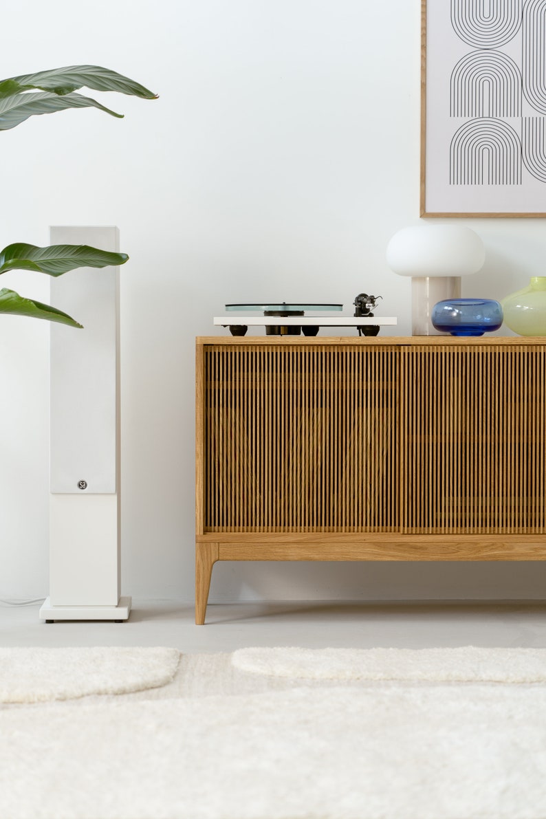 Puede incluir: Un altavoz est&eacute;reo blanco se encuentra junto a un armario de madera con un tocadiscos en la parte superior. El armario tiene un frente de listones y se apoya en cuatro patas. Una l&aacute;mpara blanca con una pantalla blanca y un jarr&oacute;n de vidrio azul est&aacute;n encima del armario. La habitaci&oacute;n tiene una pared blanca y una alfombra blanca en el suelo.