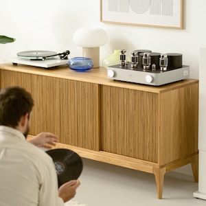 Puede incluir: Un armario de madera con dos puertas, un tocadiscos, un reproductor de discos y un amplificador vintage en la parte superior. El armario tiene un acabado de madera clara y un dise&ntilde;o acanalado en las puertas. El amplificador es plateado con botones negros y cuatro tubos de vac&iacute;o.