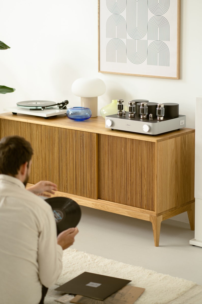 Puede incluir: Un armario de madera con dos puertas, un tocadiscos, un reproductor de discos y un amplificador vintage en la parte superior. El armario tiene un acabado de madera clara y un dise&ntilde;o acanalado en las puertas. El amplificador es plateado con botones negros y cuatro tubos de vac&iacute;o.
