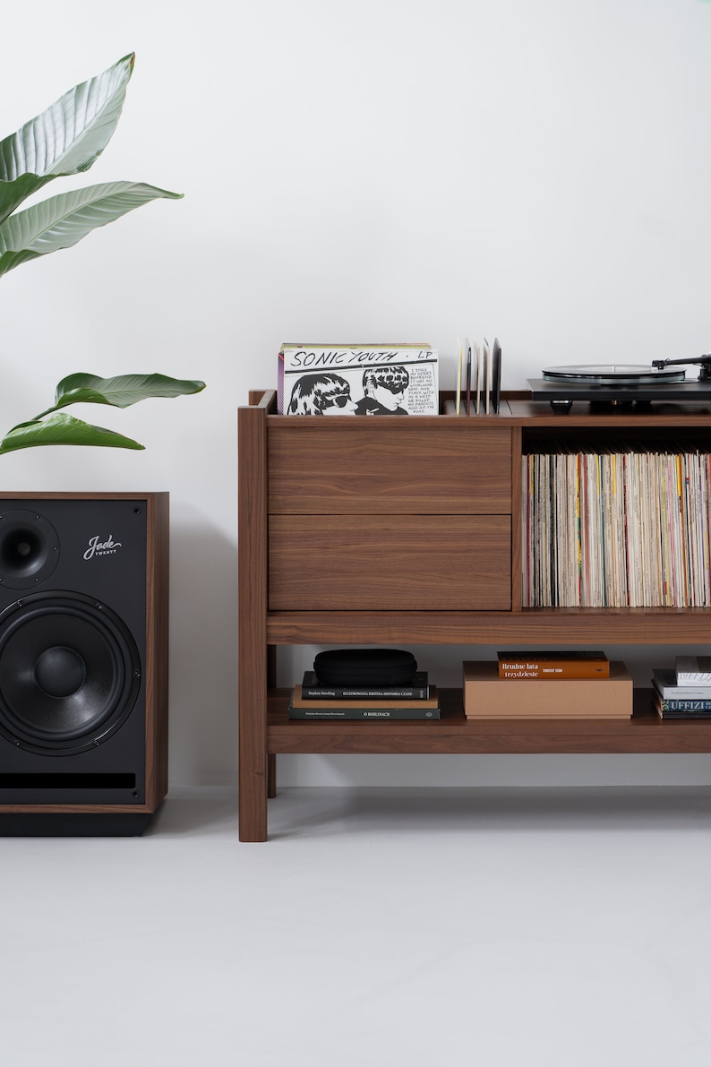 May include: A wooden record player stand with two drawers, shelves, and a turntable. The stand is brown and has a record player on top. There are records on the shelves and a speaker on the left side of the image.