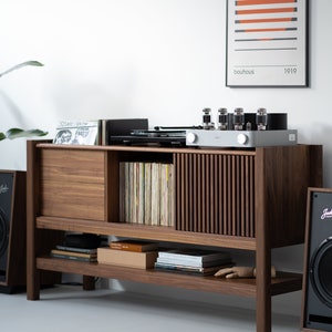 May include: A wooden record player stand with a sliding door, shelves, and a record player on top. The stand is made of dark wood and has a light wood trim. There is a vintage amplifier with vacuum tubes on top of the stand. There is a framed print with the text "bauhaus 1919" on the wall behind the stand.
