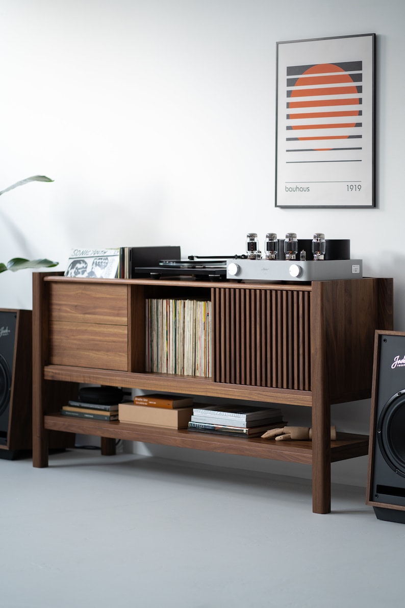 May include: A wooden record player stand with a sliding door, shelves, and a record player on top. The stand is made of dark wood and has a light wood trim. There is a vintage amplifier with vacuum tubes on top of the stand. There is a framed print with the text "bauhaus 1919" on the wall behind the stand.