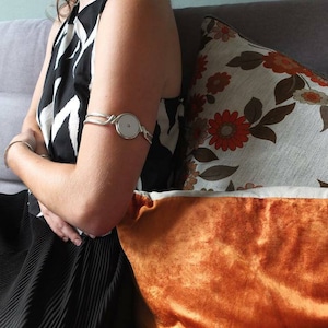 Può includere: Un bracciale d'argento con un centro circolare di colore chiaro. Il bracciale avvolge la parte superiore del braccio. Un braccialetto d'argento è visibile anche sul polso. La persona indossa un vestito a fantasia bianco e nero.