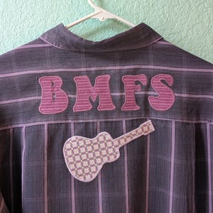 May include: A button-down shirt with a plaid pattern in shades of gray and purple. The back of the shirt features the letters "BMFS" in a decorative font, along with a guitar-shaped patch.