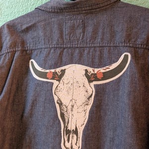 May include: A dark blue button-down shirt with a large graphic of a bull skull on the back. The skull is white with black shading and red floral accents on the horns. The shirt is hanging on a black hanger.