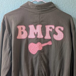 May include: Olive green jacket with the letters "BMFS" in pink, patterned fabric on the back, along with a pink guitar. The jacket is hanging on a white hanger against a light blue wall.