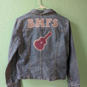 May include: A blue denim jacket with the letters "BMFS" appliqued across the upper back. Below the letters is a purple guitar applique. The jacket is hanging on a white hanger against a light green wall.