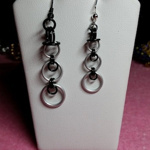 May include: A pair of silver and black dangle earrings with multiple rings. The earrings are displayed on a white stand.