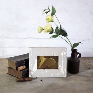 May include: A rustic still life composition. It includes a white-framed picture, two old books, a corkscrew, and a small brown vase with yellow flowers. The objects are set on a wooden table against a white brick wall.