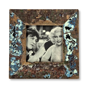 May include: A square picture frame with a distressed, weathered finish in shades of brown, blue, and light green. The frame holds a black and white photograph of two individuals, one pointing a finger while the other looks on with a smile.