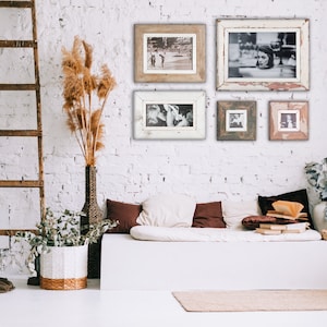 May include: A rustic interior scene featuring a white brick wall adorned with framed black and white photographs. A wooden ladder leans against the wall, and a white bench is decorated with pillows. A tall vase holds dried plants, adding a natural touch.