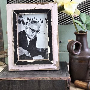 Peut inclure: Une photographie vintage en noir et blanc dans un cadre vieilli rose clair et noir. La photographie présente un homme portant des lunettes et un costume. Un vase marron avec une poignée et une fleur jaune sont également dans l'image.
