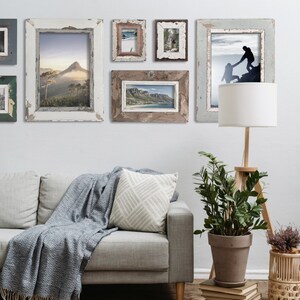 May include: A living room scene featuring a gray sofa with a blue blanket and patterned pillow. Several framed landscape photos hang on the wall. A wooden floor lamp and potted plant add to the decor. The frames have a distressed, rustic look.