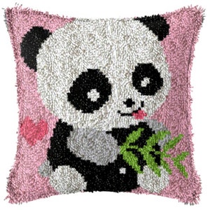 Puede incluir: Un cojín cuadrado hecho a mano con un diseño de oso panda. El panda tiene pelaje blanco y negro, una lengua rosa y sostiene hojas de bambú verdes. El cojín tiene un borde rosa y un corazón rosa.