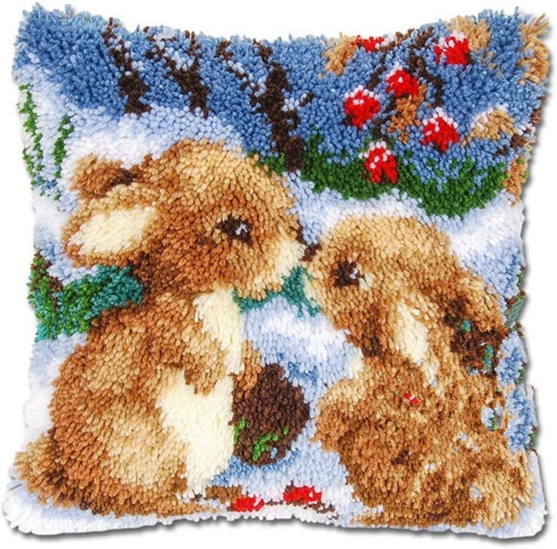 May include: A close-up of a needlepoint pillow featuring two brown rabbits in a snowy winter scene. The rabbits are looking at each other and there are red berries in the background.