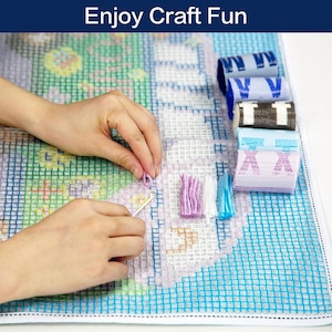May include: A person is working on a colourful needlepoint project. The project is on a white gridded canvas with a blue border. The person is using a needle and yarn to create a design. There are several spools of yarn in different colours on the right side of the image.