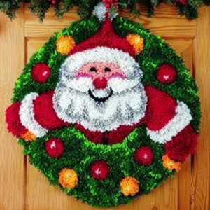 May include: A Christmas wreath featuring Santa Claus. The wreath is circular, with a green base and red, white, and orange accents. Santa has a white beard, red hat, and rosy cheeks. The wreath is decorated with red and orange ornaments.