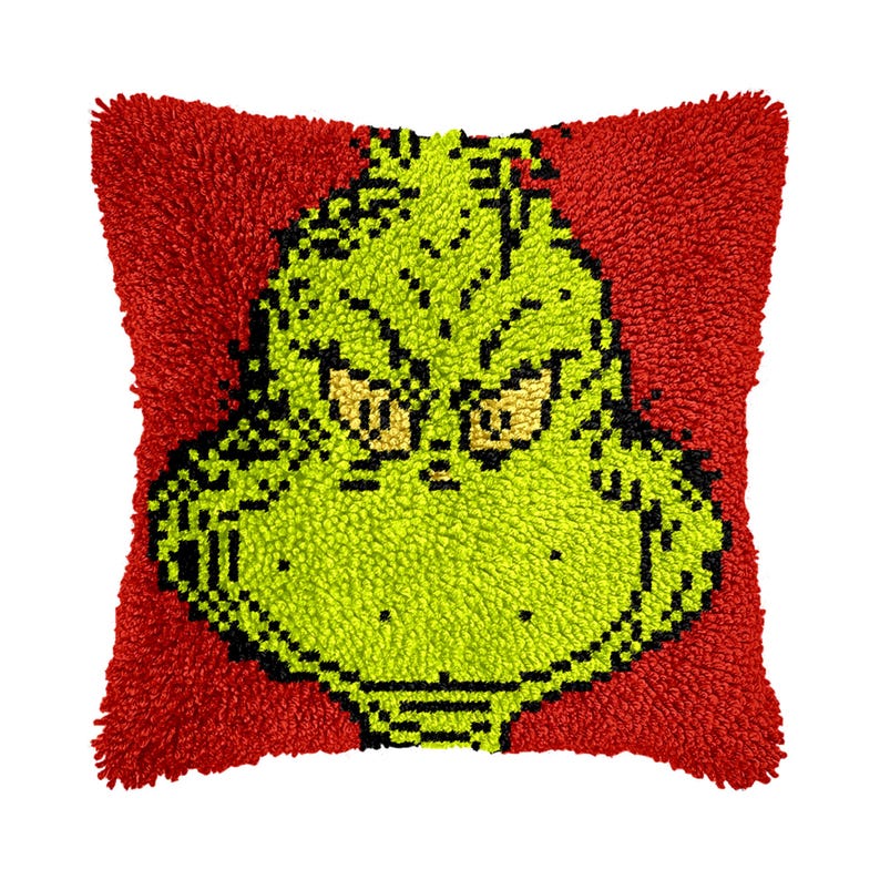 May include: A green Grinch face with a grumpy expression is embroidered on a red plush cushion.