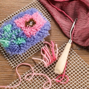 May include: A colorful punch needle project with a floral design, featuring pink, blue, green, and brown yarn. A wooden-handled punch needle tool and pink yarn are also visible, along with a white grid canvas. The craft supplies are arranged on a wooden surface.