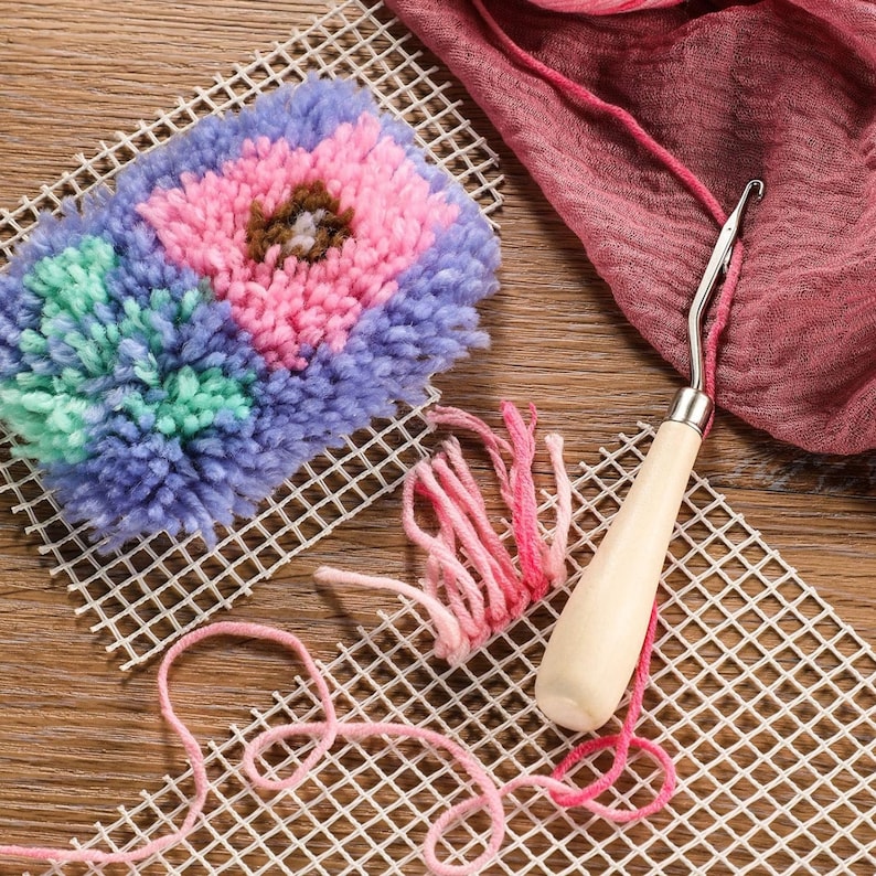 May include: A colorful punch needle project with a floral design, featuring pink, blue, green, and brown yarn. A wooden-handled punch needle tool and pink yarn are also visible, along with a white grid canvas. The craft supplies are arranged on a wooden surface.