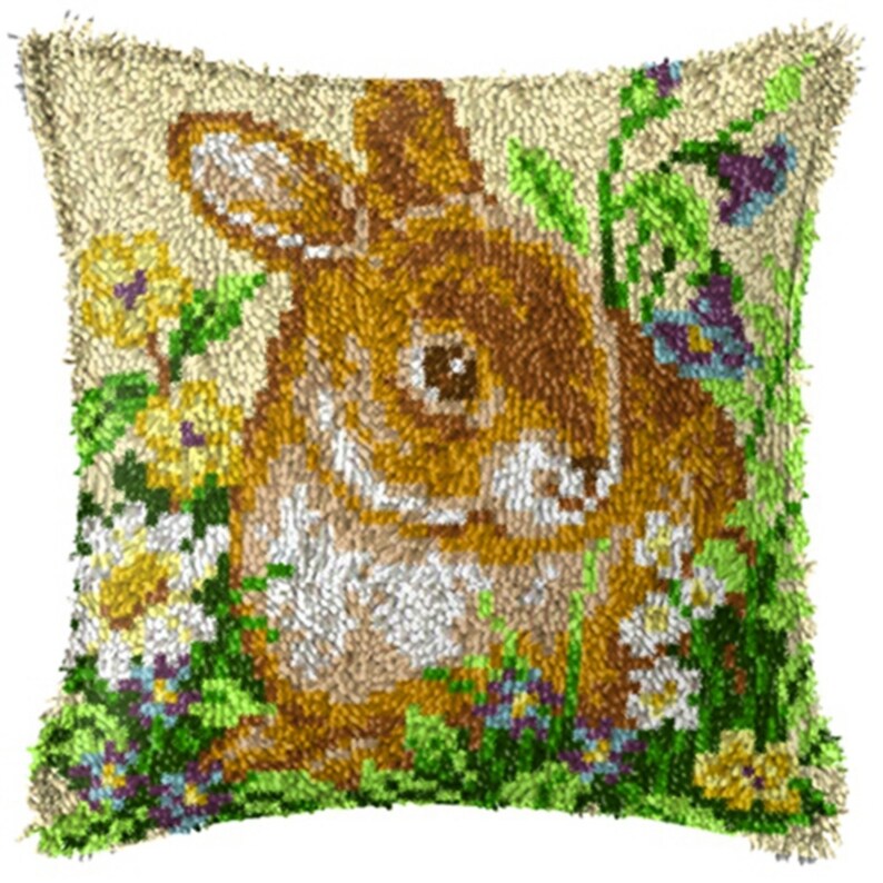 May include: A beige pillow with a brown bunny rabbit surrounded by green leaves and colorful flowers. The bunny has brown fur, a pink nose, and black eyes. The flowers are yellow, white, and purple.