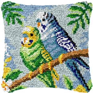 以下が含まれることがあります： 茶色の枝に止まっている2羽のセキセイインコの詳細なニードルポイントデザインが施された正方形の装飾枕。鳥は緑、黄色、青の色合いで、緑の葉のある水色の背景に設定されています。