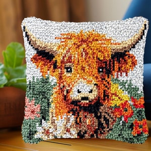 May include: Square decorative pillow featuring a detailed, textured image of a Highland cow. The cow has orange and brown fur, large horns, and is set against a background of green and red flowers. The pillow has a light gray border.