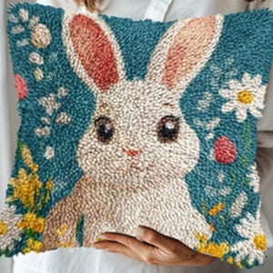 Könnte beinhalten: Ein quadratisches, strukturiertes Kissen mit einem weißen Hasen mit rosa gefütterten Ohren und dunklen Augen, vor einem tealblauen Hintergrund. Das Design umfasst florale Akzente in Gelb, Weiß und Rosa, wodurch ein charmantes, dekoratives Stück entsteht.