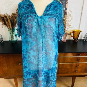 Op de afbeelding: Een knielange jurk in blauwtinten met een bloemenpatroon. De jurk heeft korte mouwen en een V-hals met een trekkoord. De stof is doorschijnend en licht. De jurk wordt op een paspop gepresenteerd.