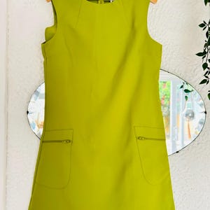 May include: A sleeveless, lime green dress with two front pockets and a zipper closure. The dress is hanging on a wooden hanger against a white wall with a decorative mirror in the background. The dress has a simple, classic design.