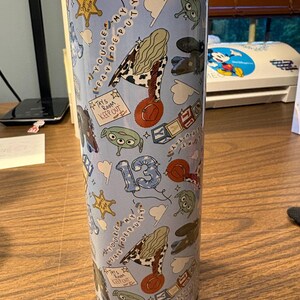 May include: A cylindrical tumbler with a light blue background featuring a repeating pattern of cartoon aliens, sheriff badges, cowboy hats, alphabet blocks, and the number 13. Text includes "You're my favorite deputy" and "Tay's room keep out".