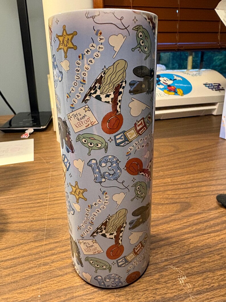 May include: A cylindrical tumbler with a light blue background featuring a repeating pattern of cartoon aliens, sheriff badges, cowboy hats, alphabet blocks, and the number 13. Text includes "You're my favorite deputy" and "Tay's room keep out".