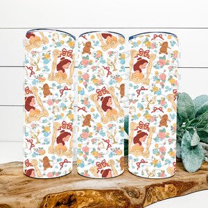 May include: Three white insulated tumblers with a repeating pattern of a portrait of a woman in a gold frame, red bows, and floral elements. The tumblers are decorated with a design featuring a cartoon character and floral elements.