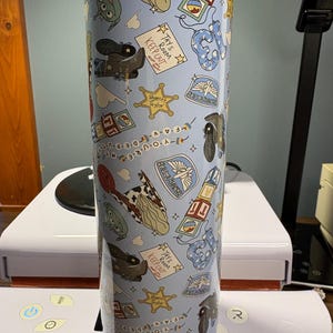May include: A cylindrical tumbler with a light blue background featuring a repeating pattern of cartoon motifs including cowboy boots, sheriff badges, alphabet blocks, alien characters, and signs that read Tay's Room Keep Out.