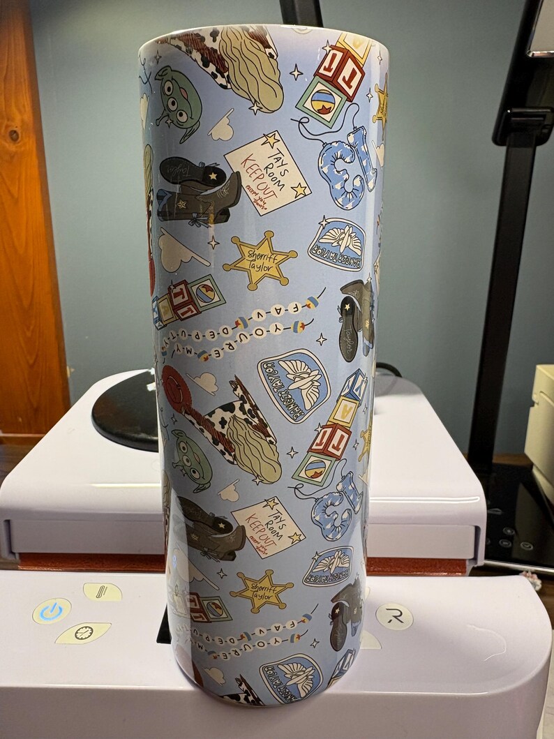 May include: A cylindrical tumbler with a light blue background featuring a repeating pattern of cartoon motifs including cowboy boots, sheriff badges, alphabet blocks, alien characters, and signs that read Tay's Room Keep Out.
