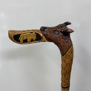 Peut inclure: Une canne de marche en bois avec une poignée sculptée en tête de bison. La poignée est brune et présente un motif tissé. La canne est en bois clair et a une teinte brune.
