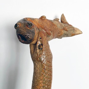 Peut inclure: Une canne de marche en bois sculptée en forme de serpent avec un poisson dans sa gueule. Le serpent est brun et vert avec une surface texturée. Le poisson est brun et orange avec un visage détaillé.