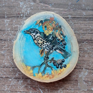 Peut inclure: Une tranche de bois peinte à la main représentant un oiseau perché sur une branche avec des baies. L'oiseau est peint dans des tons de brun, noir et blanc, sur un fond bleu. La branche est peinte en brun et vert, avec des feuilles jaunes et des baies noires.