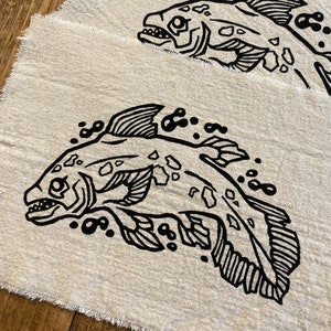 May include: Two beige linen squares featuring a black fish illustration. The fish is depicted with detailed fins, scales, and an open mouth. Bubbles surround the fish, and the linen edges are frayed.