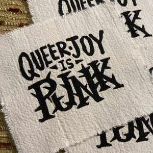May include: Close-up of three off-white fabric patches with black text. The central patch reads "QUEER JOY IS PUNK" in a bold, stylized font. The other patches display fragments of the same message. The edges of the patches are frayed.