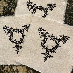 Könnte beinhalten: Drei cremefarbene Stoffflicken mit einem schwarzen Transgender-Symbol-Design. Das Symbol ist ein Kreis mit Pfeilen, die nach oben und zu den Seiten zeigen, und einem Kreuz unten, alles umgeben von Dornen.