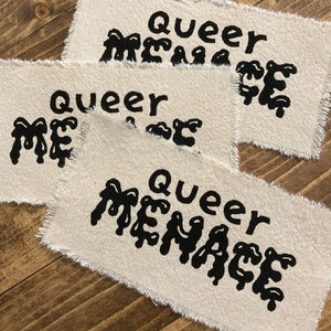 Può includere: Tre toppe di tessuto con la frase "Queer Menace" stampata in lettere nere che gocciolano su uno sfondo beige.