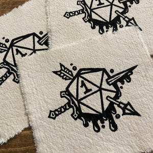 Può includere: Sottobicchieri quadrati in tessuto bianco sporco con una grafica nera di un dado D20 trafitto da una spada e una freccia. Il dado mostra il numero 1. I bordi dei sottobicchieri sono sfilacciati.
