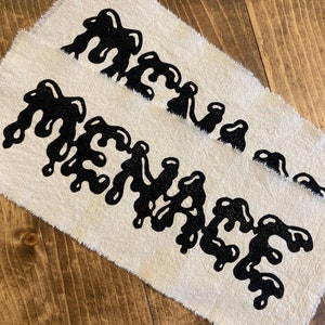 May include: Two white fabric patches with black dripping text that reads "MENACE".