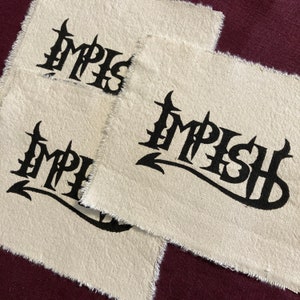 May include: Three beige fabric patches with the word "IMPISH" printed in black, gothic-style lettering. The patches have a raw, frayed edge. The patches are arranged on a dark burgundy surface.
