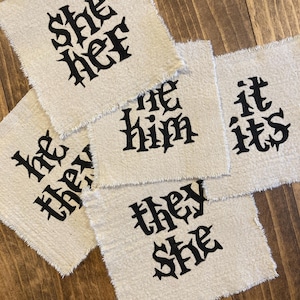 Puede incluir: Cuatro cuadrados de tela beige con texto bordado en negro. El texto en los cuadrados dice "she her", "he him", "it its" y "they she".