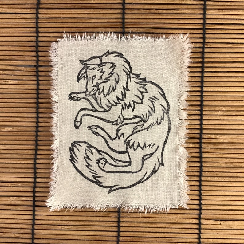 Może przedstawiać: Kwadratowa, biała naszywka z czarną ilustracją stylizowanego wilka. Wilk jest zwinięty, a jego ogon dotyka głowy. Krawędzie naszywki są postrzępione. Tło to bambusowa roleta.