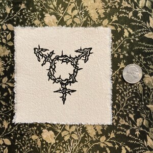 K&ouml;nnte beinhalten: Quadratischer, cremefarbener Stoffflicken mit einem schwarzen Transgender-Symbol-Design. Das Symbol ist ein Kreis mit einer Dornenkrone und Pfeilen. Der Flicken hat einen rohen, ausgefransten Rand. Eine M&uuml;nze liegt daneben.