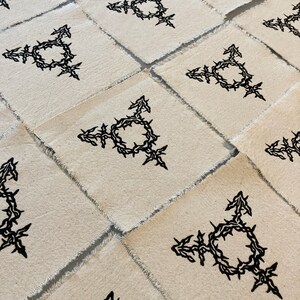 K&ouml;nnte beinhalten: Mehrere quadratische Stoffflicken mit rohen Kanten, die jeweils ein schwarzes Transgender-Symbol zeigen. Das Symbol ist eine Kombination aus m&auml;nnlichen und weiblichen Symbolen und einem Kreis, der mit einem dornigen Rankenmuster verflochten ist.